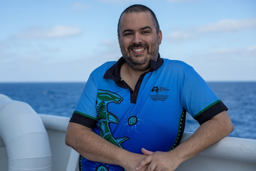 A man in a blue t-shirt smiles while leaning on the rail of a boat with the ocean in the background.