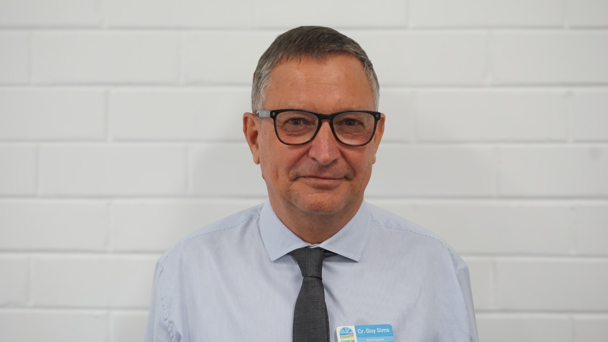 Un hombre de pelo corto y oscuro se encuentra frente a una pared clara. Lleva una camisa abotonada y sonríe.