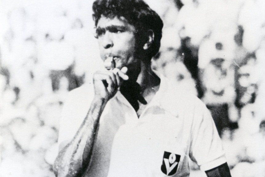 a black and white photo of an indigenous man umpiring a game of AFL, he is blowing a whisle