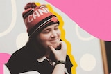 Shoshana smiles and scratches her chin in a beanie, surrounded by colourful graphics.