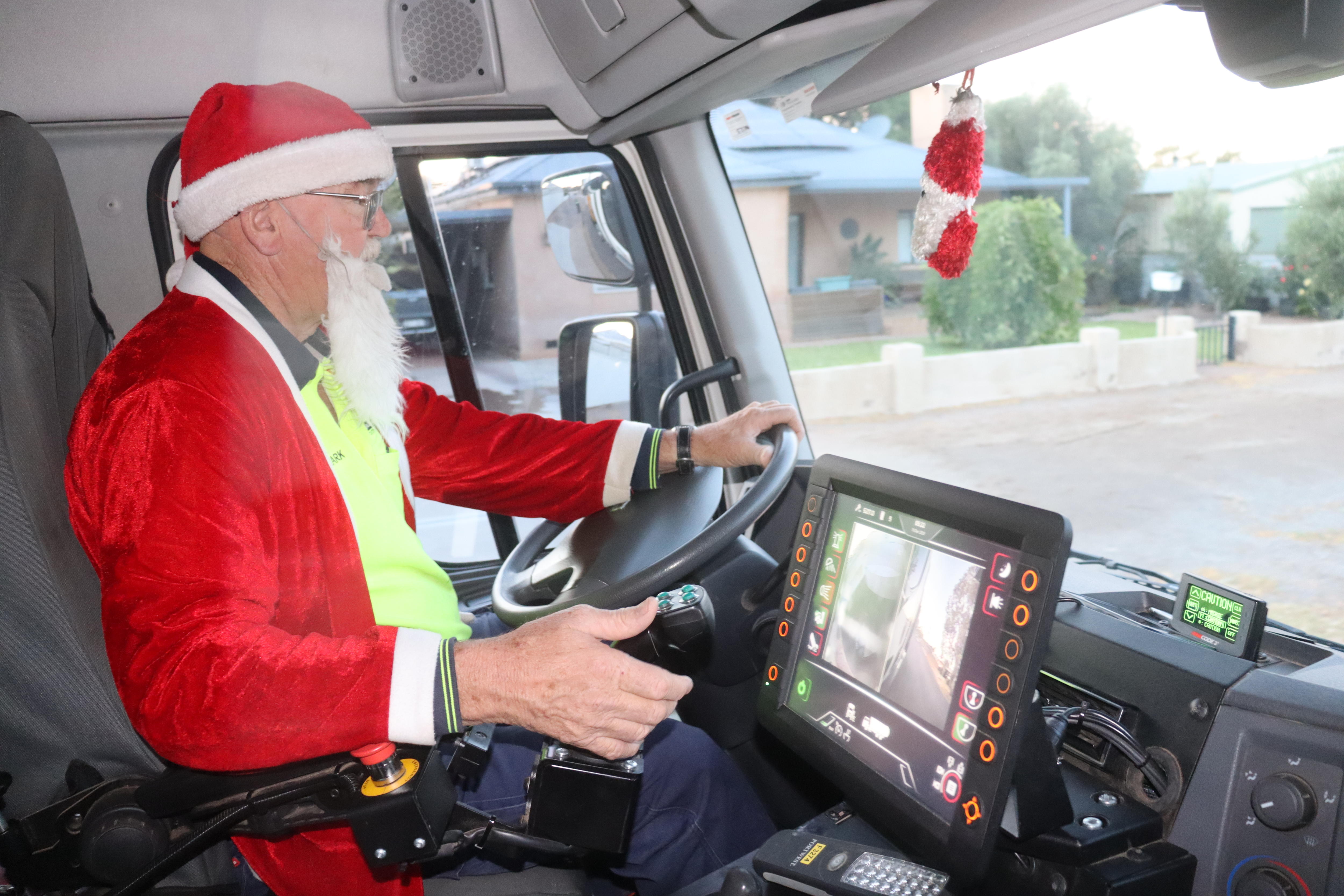 Un hombre mayor con un top de neón y disfraz de Papá Noel en un camión de basura, con una mano en un volante y la otra en un joystick.
