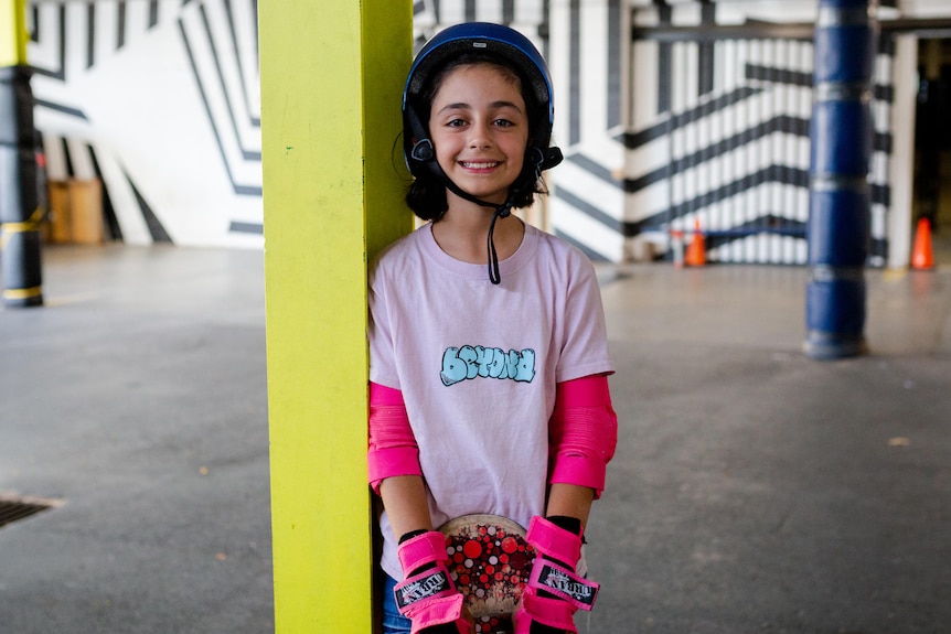 A bright young girl in a pink shirt leans against a pole, holding her skateboard.
