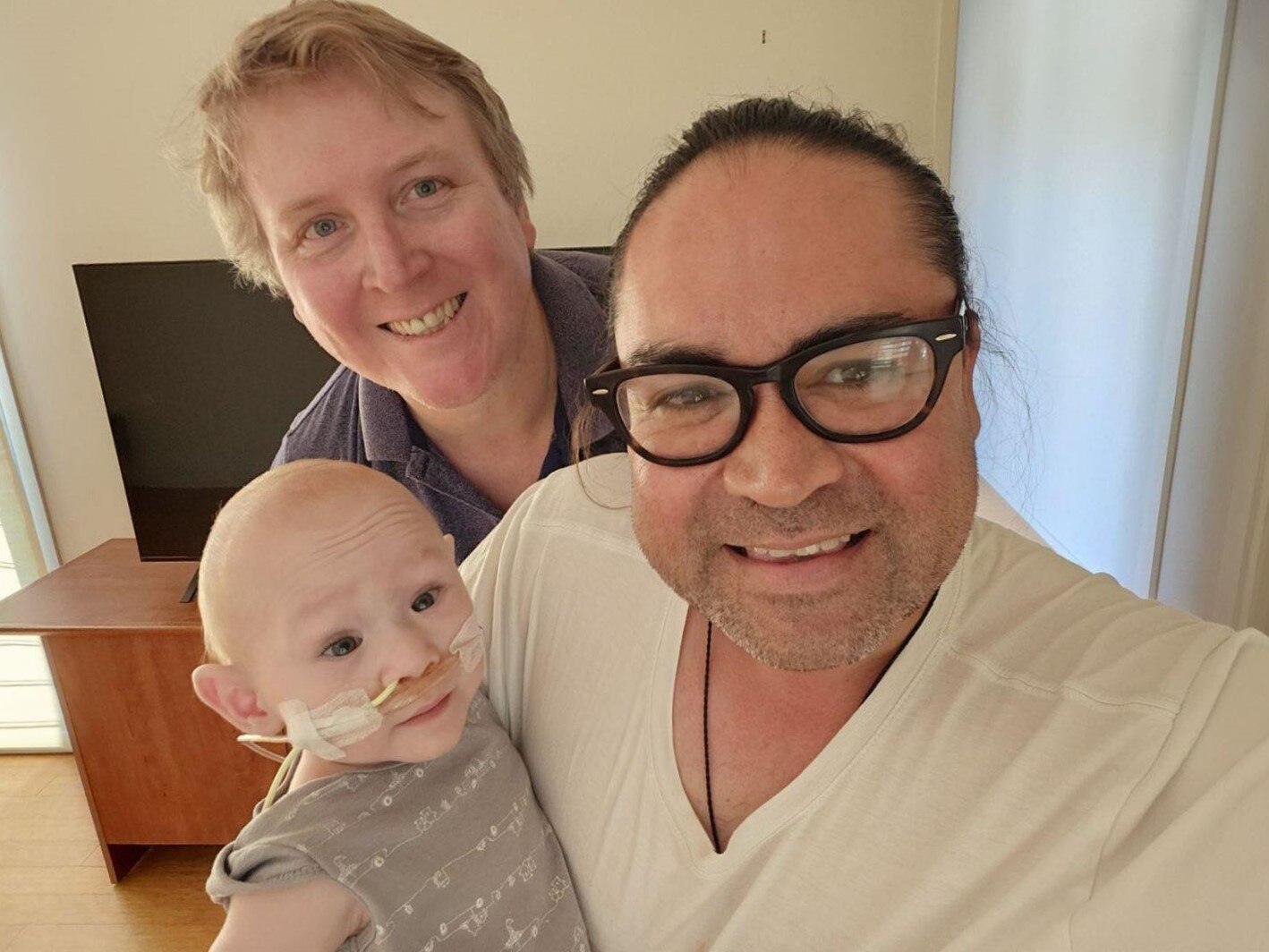 A selfie man with his partner and baby son who has a feeding tube all smiling into the camera