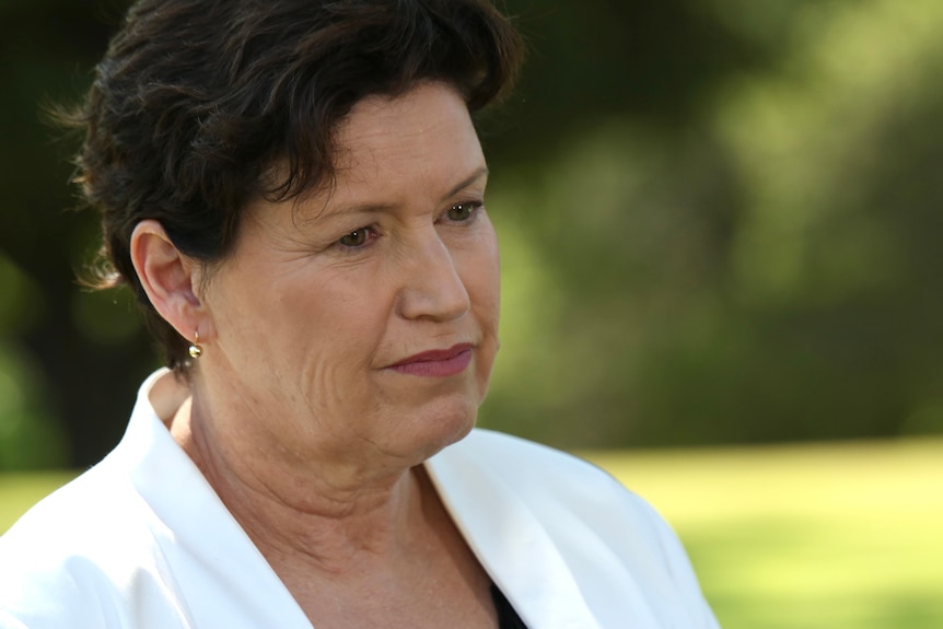 A dark-haired woman in a white suit looks serious. 