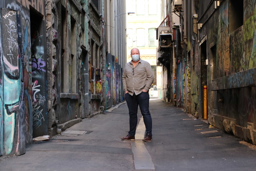 A man wearing a face mask stands in an alley with his hands in his pockets.