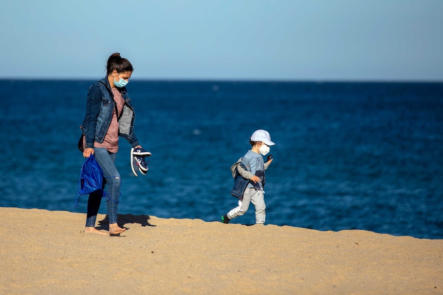 A woman and her son wear face masks as they walk on a beach.