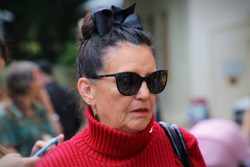 Une femme d'âge moyen portant des lunettes de soleil, un pull rouge et un nœud noir dans les cheveux.