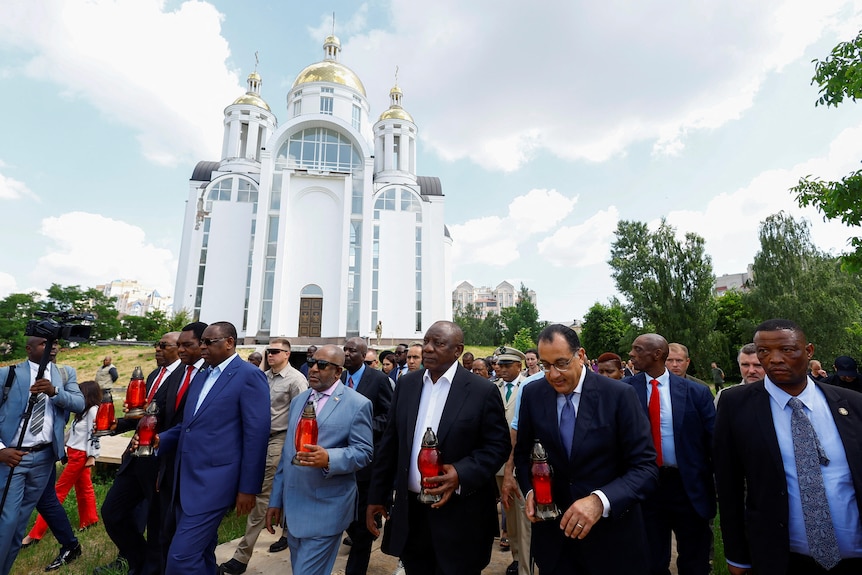 A large white church is pictured with men in suits walking in front of it carrying candles.