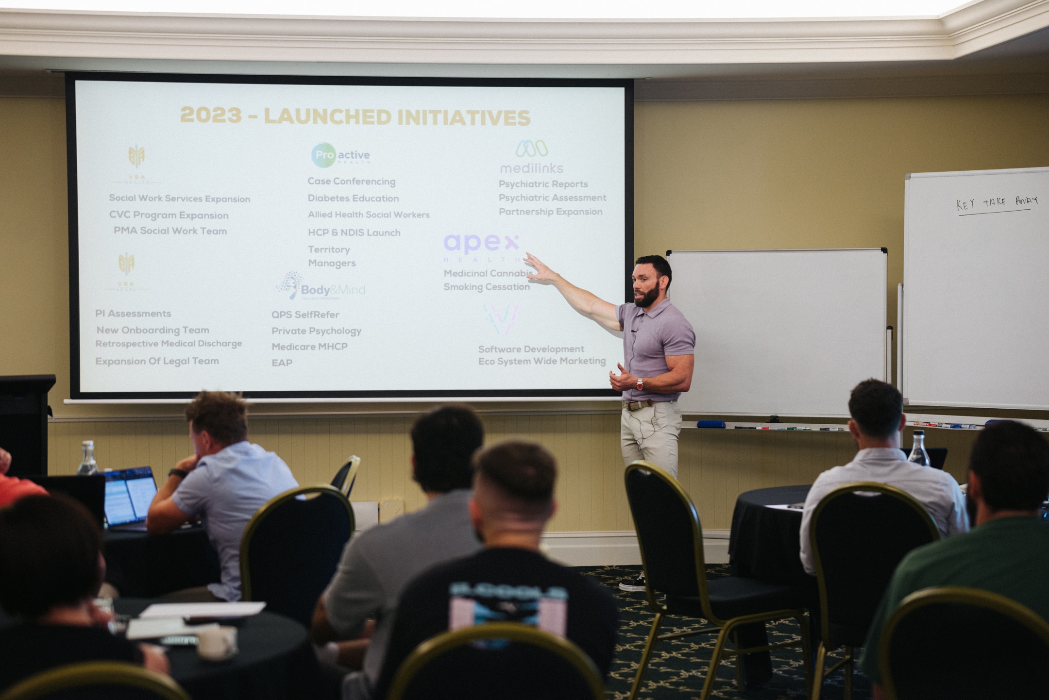 A young man wearing a purple collared shirt and chinos pointing to a powerpoint presentation.