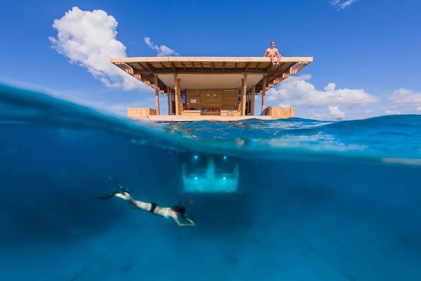 A man sits on the roof of a room floating in the ocean. A woman swims beneath the surface, near the submerged bedroom.