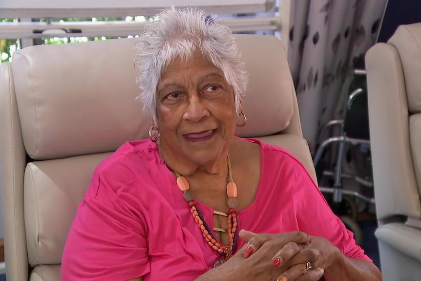 a woman sitting in an armchair wearing a bright pink t shirt and colourful necklaces