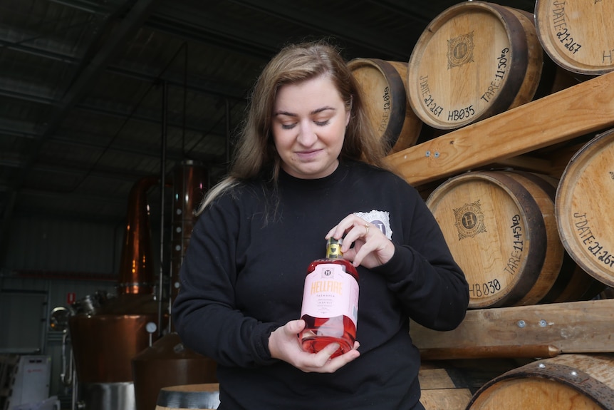 A woman standing in front of wooden barrels looks down at a bottle of non-alcoholic spirits