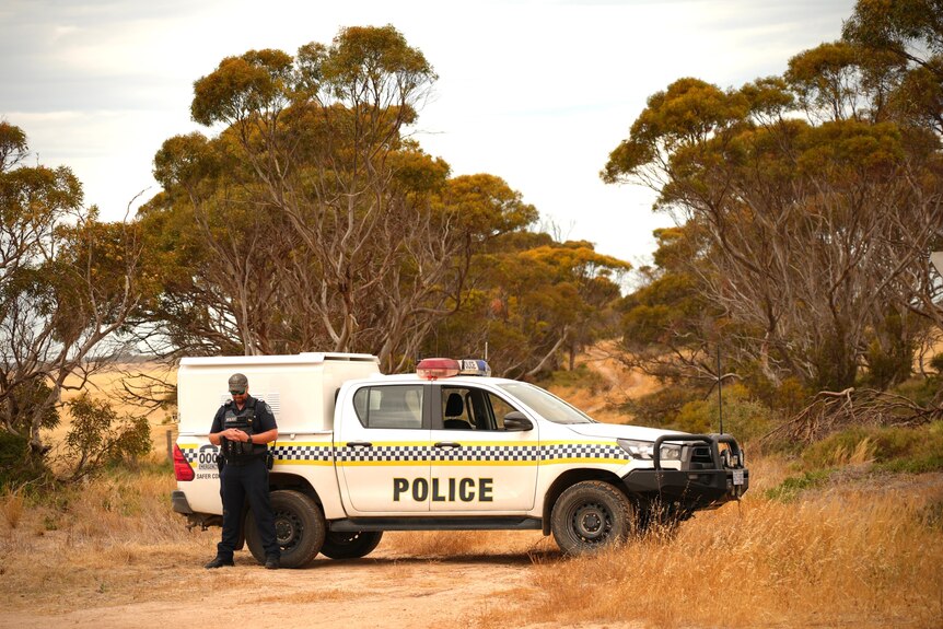 A police officer stands up against a police vehicle parked on a dirt road