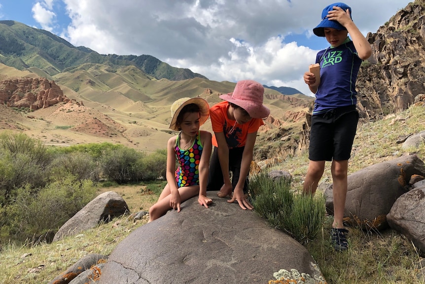 Three children look at a rock with a small engraving on it, with hills in the background 