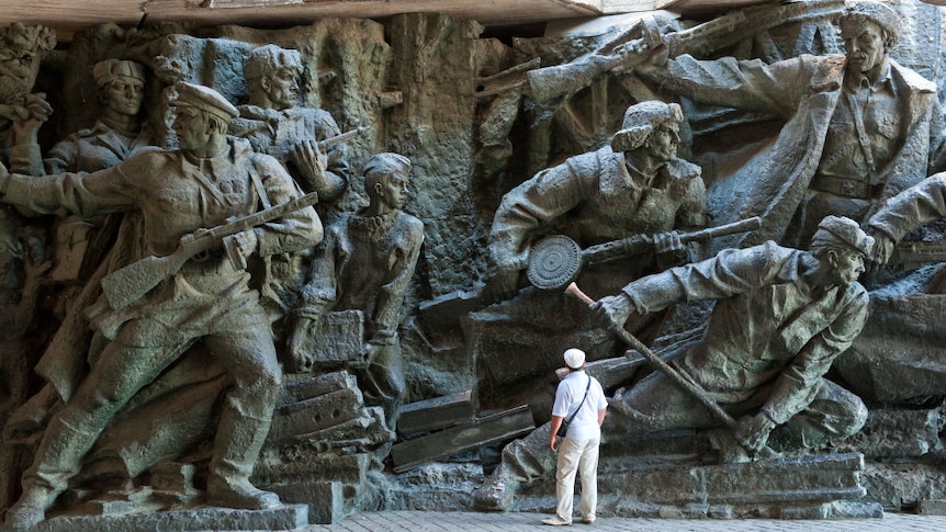 A man stands in front of a large sculpture scene depicting the Patriotic War of 1941-1945