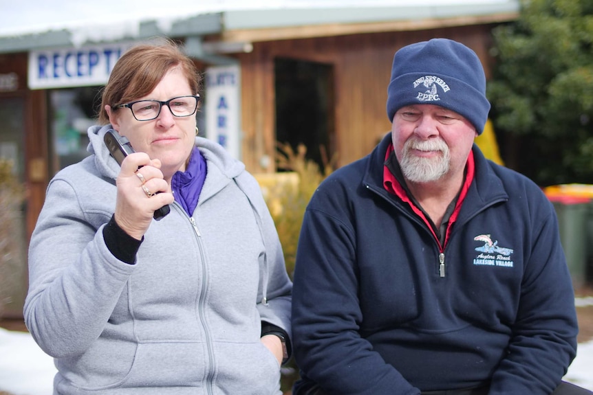 A woman holds a telephone up while sitting next to a man wearing a beanie and jumper. There is snow on the ground.