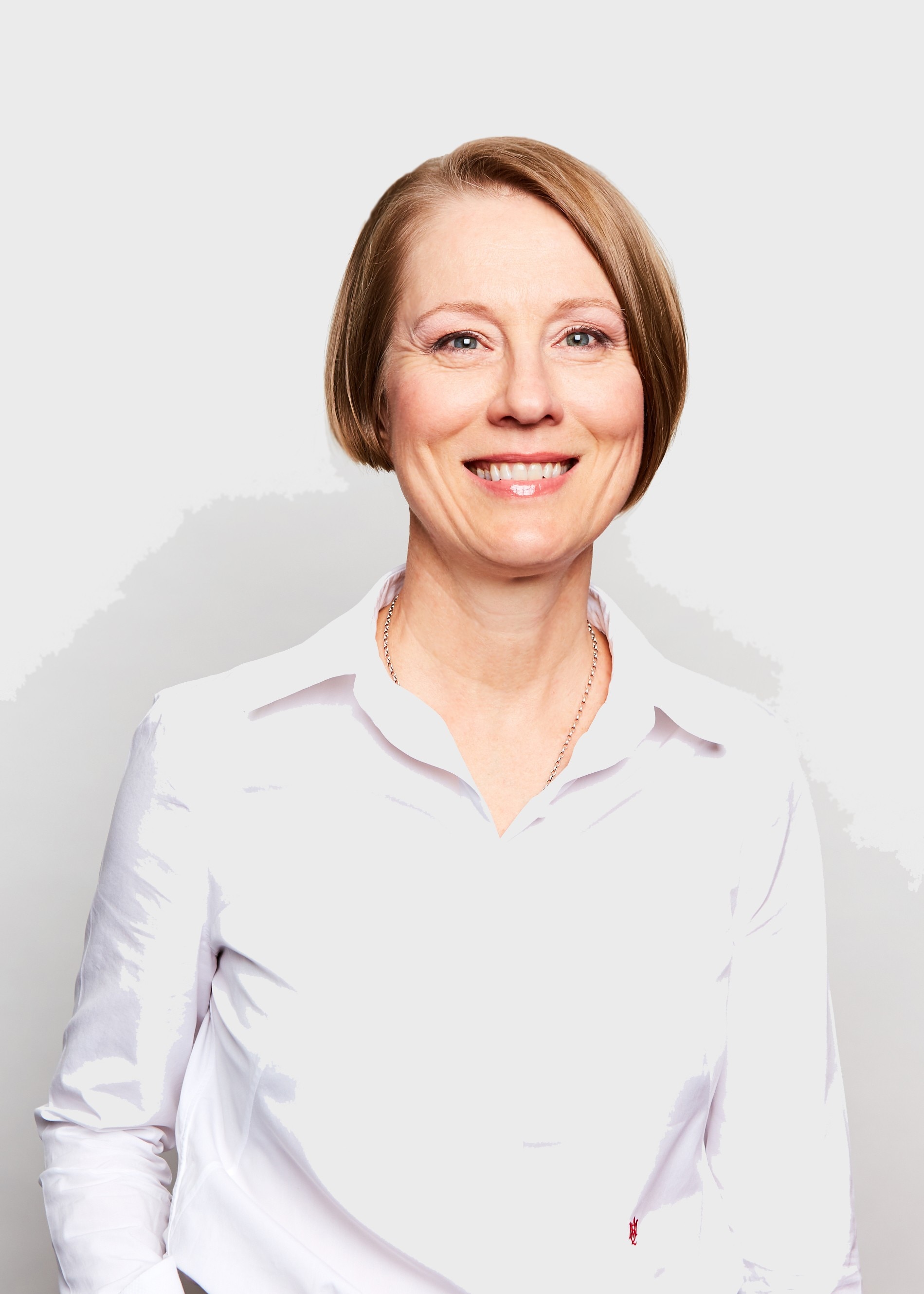 Una mujer con el pelo rojo hasta la mandíbula y raya al lado sonríe a la cámara. Lleva una camisa de cuello blanca y está parada frente a una pared blanca.