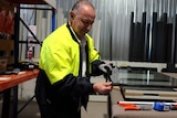 A balding man in a hi-vis jacket places a rubber trim around a plane of glass