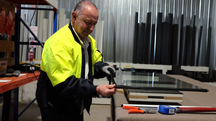 A balding man in a hi-vis jacket places a rubber trim around a plane of glass