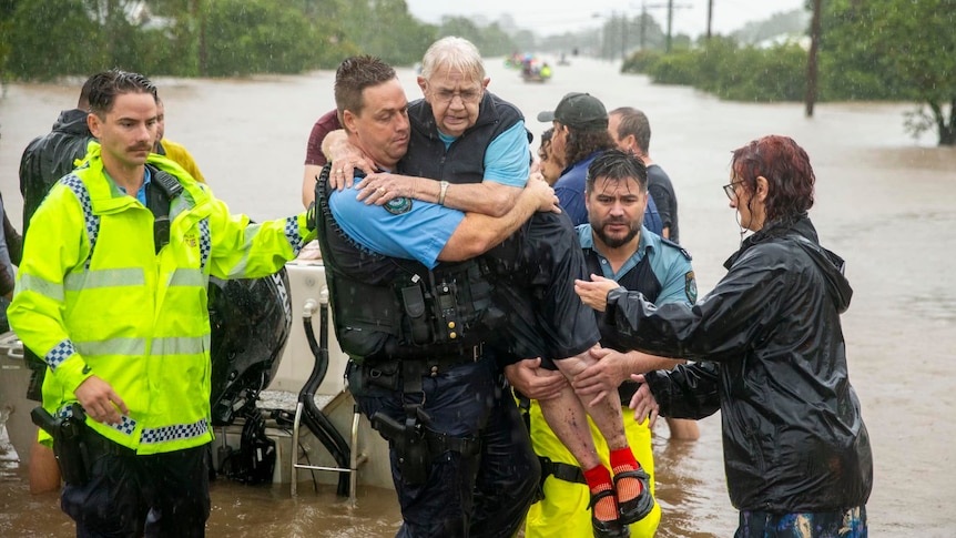 An elderly woman is soaked in rain and being carried by police officers through flood water