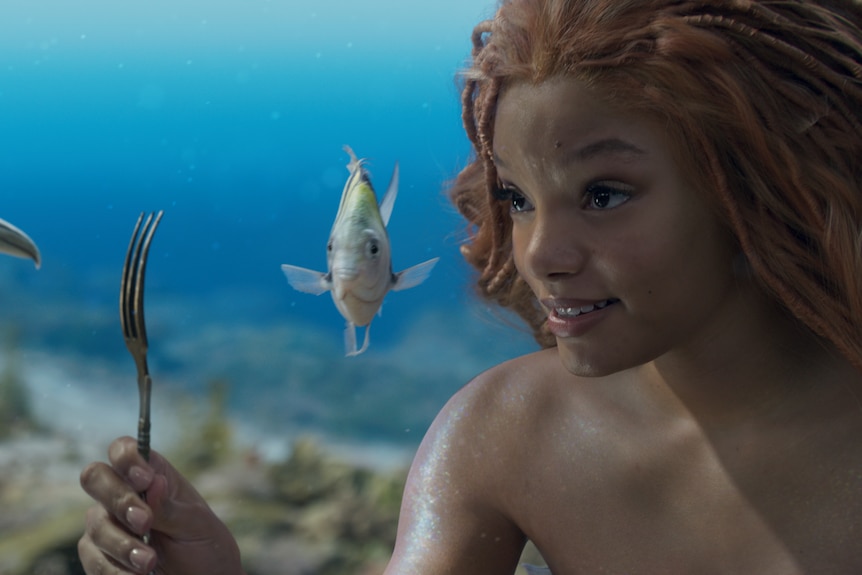 A young black woman with red-toned dreadlocked hair is pictured under the sea, holding a fork. A flounder fish is at her side.