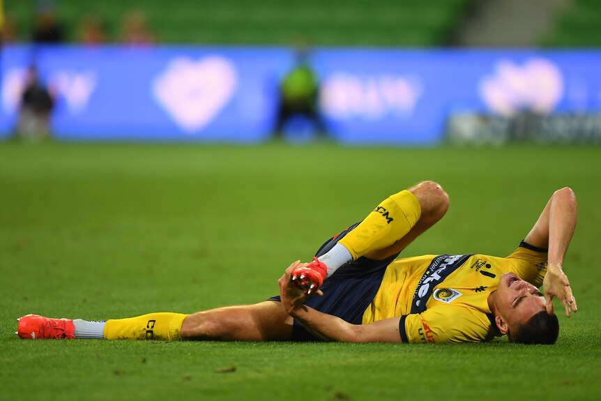 An A-League men's player lies on the ground clutching his foot in pain during a game.