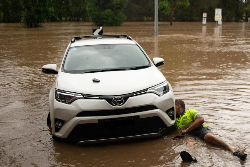 A man lies in floodwater beside a stranded car, working near the tyre.