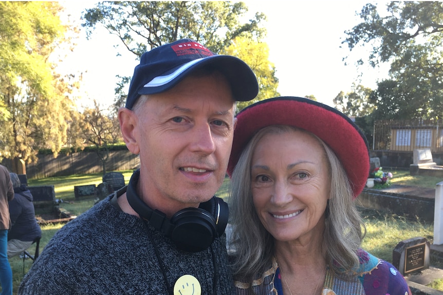 A man with a blue cap and headphones around his neck smiles next to a lady in a red hat and a multi-colour blouse.