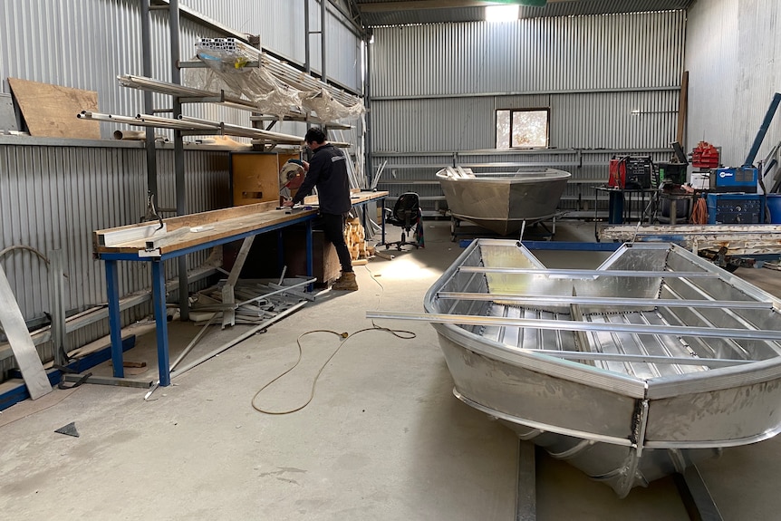 A man is standing in a shed, cutting metal. There is a boat in the front. 