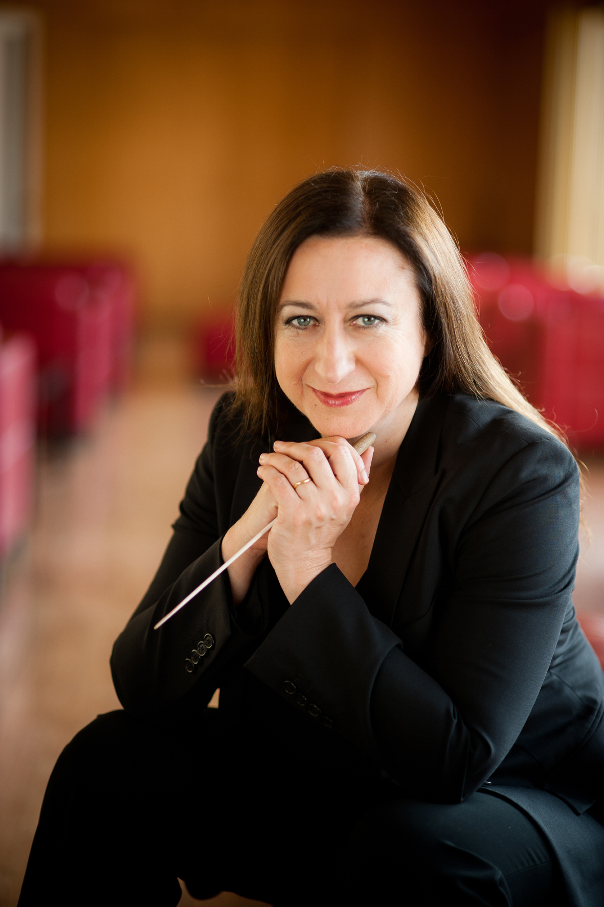 White middle-aged woman with long brown hair wears a black suit and holds a conductor's baton.