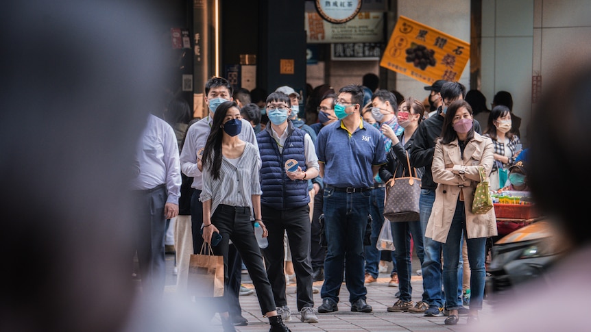 A group of people in masks stand at a street corner, waiting to cross, in Taipei
