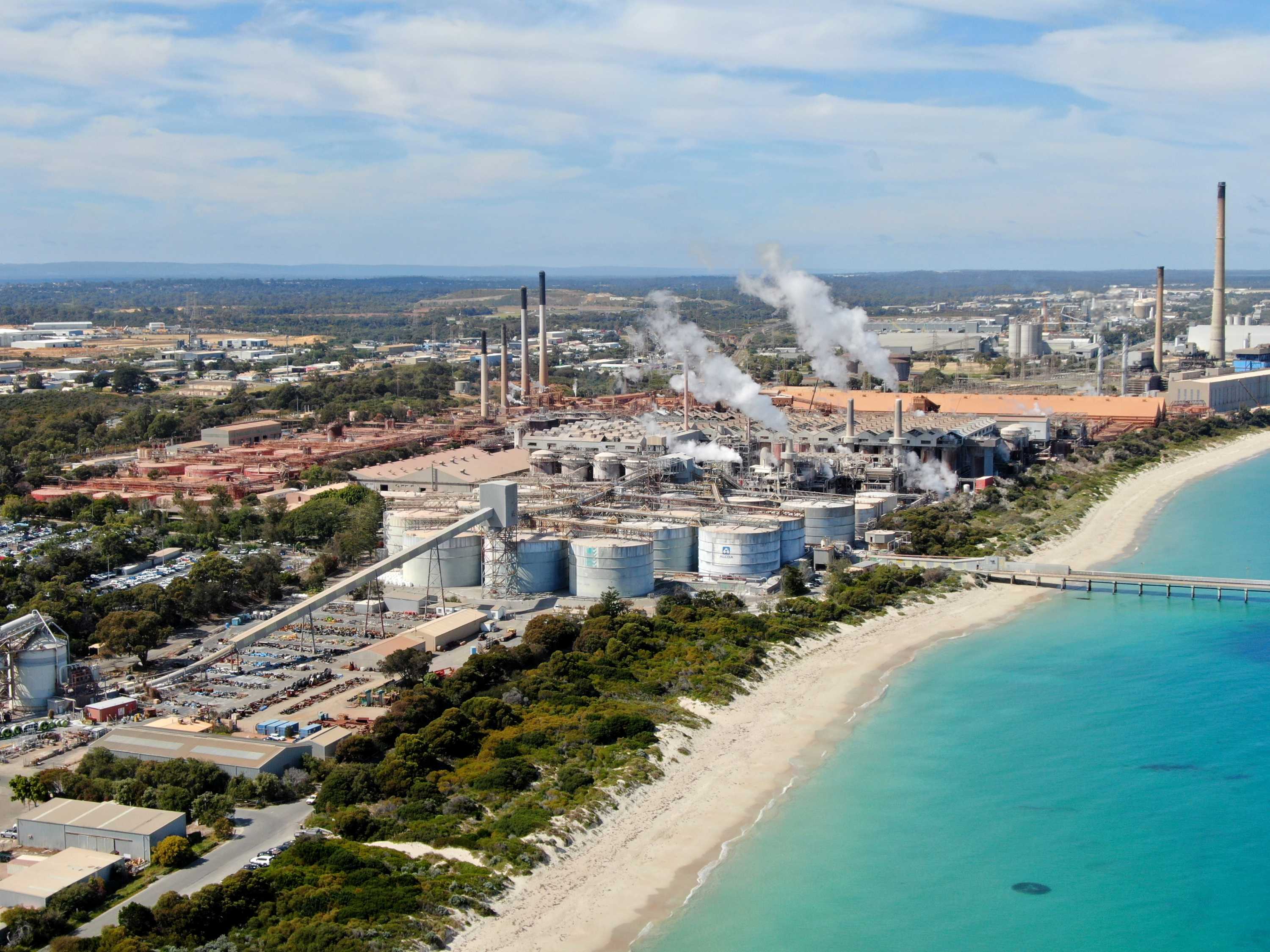 An empty beach leads to a heavy industrial area with large smoke plumes