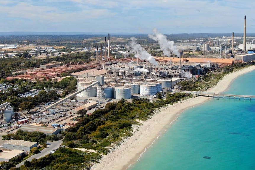 An empty beach leads to a heavy industrial area with large smoke plumes