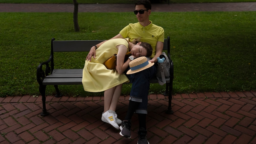 A woman wearing a sundress naps on the lap of a man wearing sunglasses as they sit on a park bench
