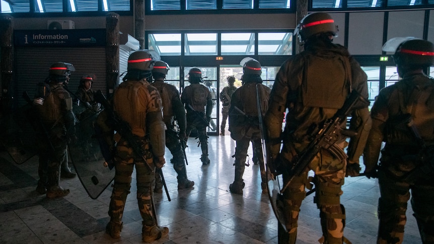 Nine soldiers stand inside in uniform wearing helmets, while others carry riot shields looking away from the camera