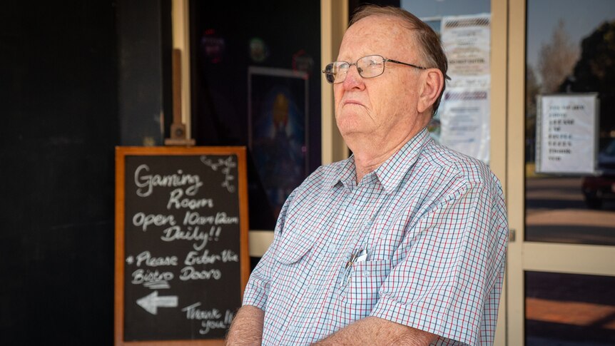 Older man wearing glasses stands arms folded outside building entrance with handwritten sign for 'gaming room' visible
