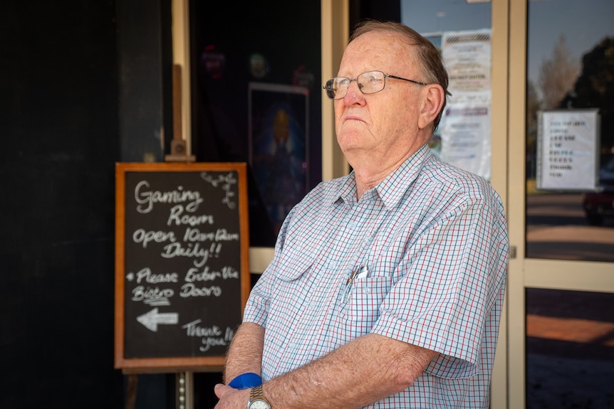 Older man wearing glasses stands arms folded outside building entrance with handwritten sign for 'gaming room' visible