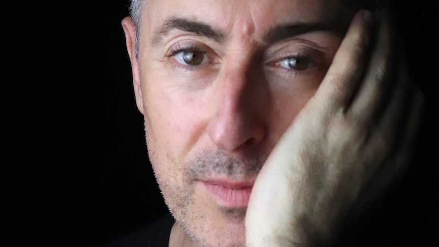 A portrait of Alan Cumming in front of a black backdrop resting his head on his hand.
