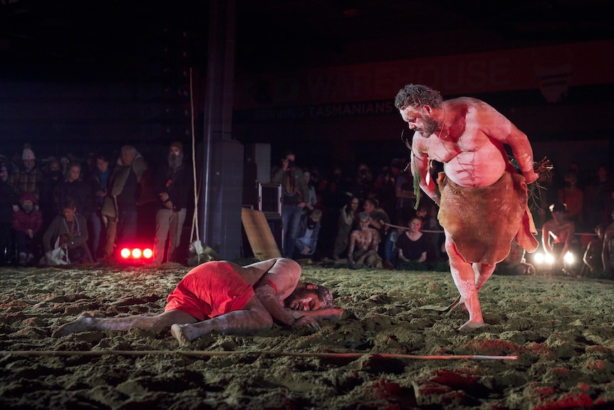 Two Aboriginal performers in body paint, one lying on the ground, while the other looks down at him, arms at his sides