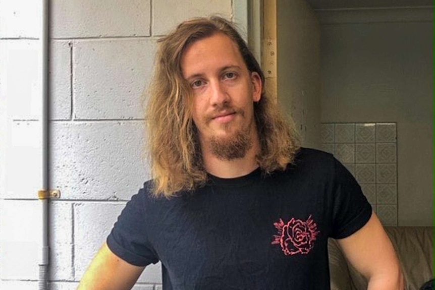 Hugh Scott standing in a black t-shirt, he has long hair and a beard.