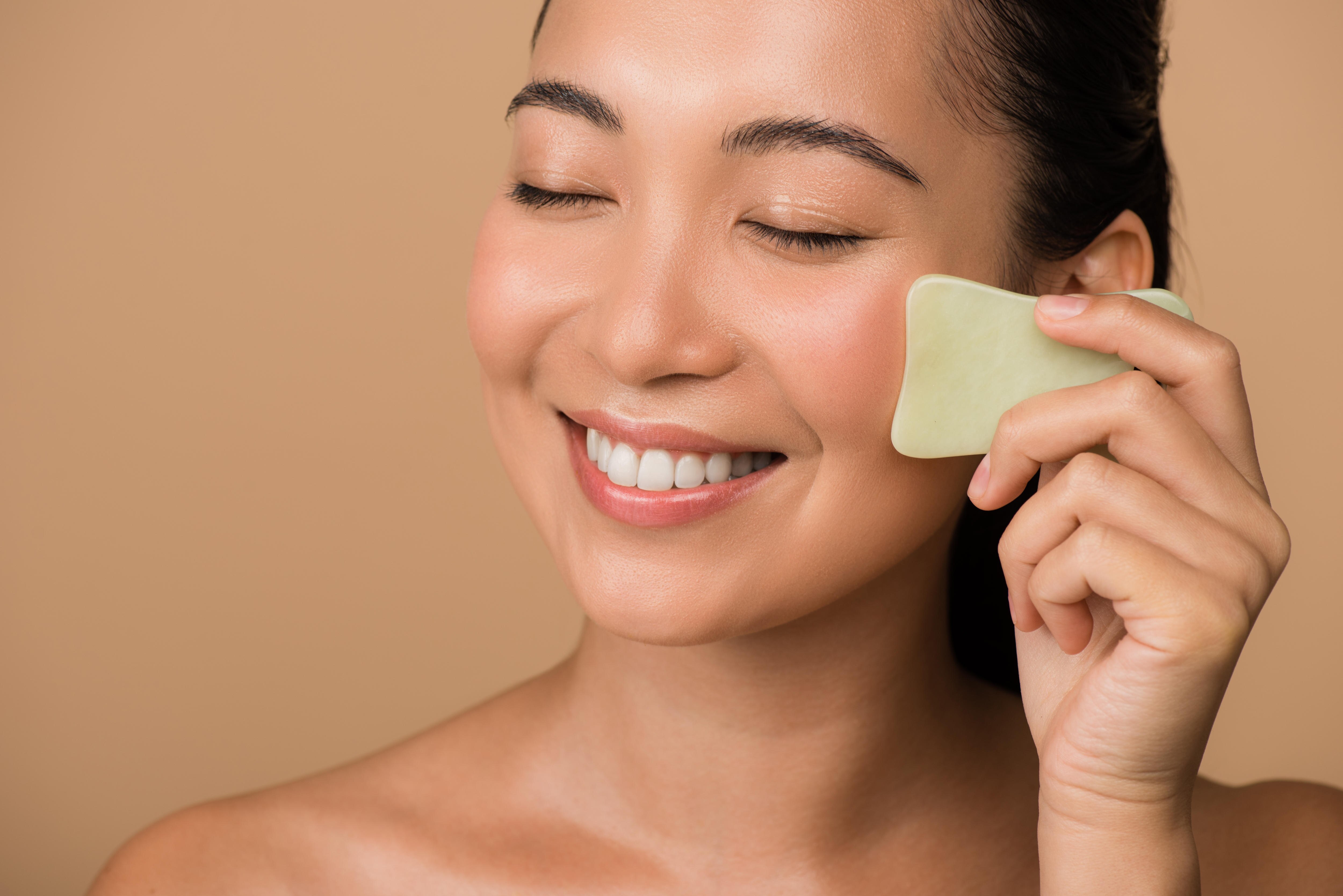 An Asian woman smiles and does gua sha on her face with a tool.