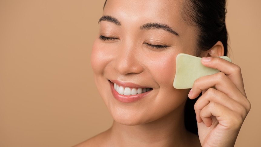 An Asian woman smiles and does gua sha on her face with a tool.