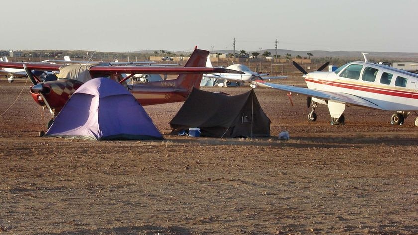 Two light planes parked in a dirt field with tents erected under their wings.