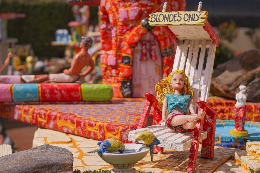 A barbie doll lounges on a small wooden deckchair outside with the words 'blondes only' above it.