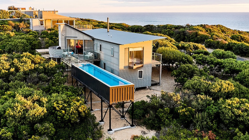 Ariel shot of large steel house on hill with a shipping container pool off the back veranda on large steel stilts. 