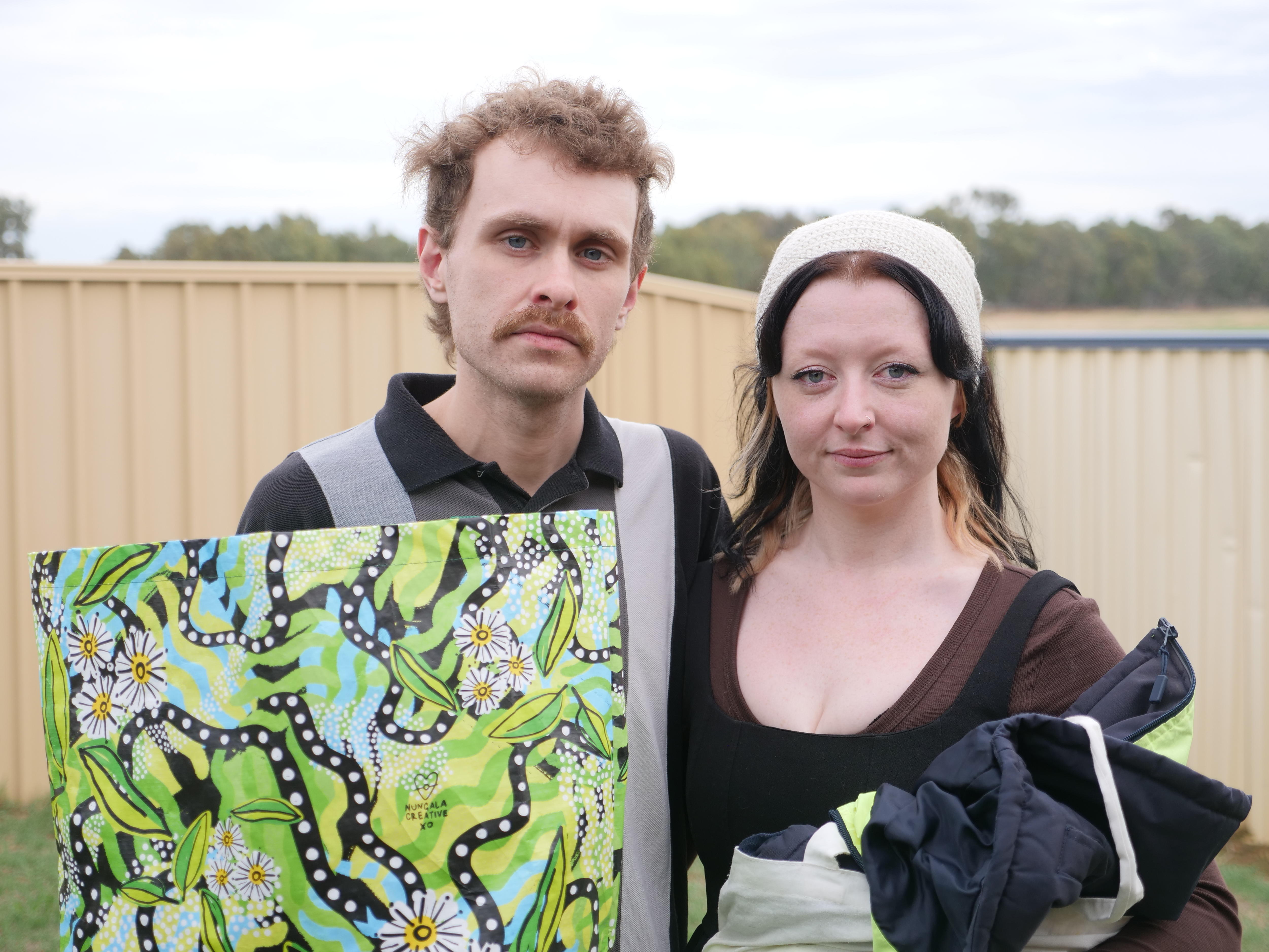 A man and a woman look seriously at the camera, outside under and overcast sky.