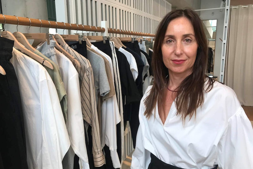 Bassike co-founder Deborah Sams stands beside a rack of clothes in a room.