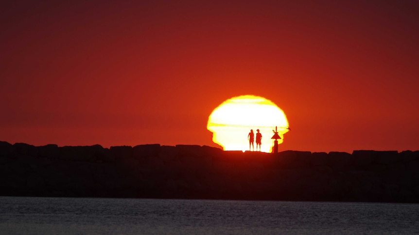 Two people silhouetted by a blazing yellow sun against a firey red backdrop.