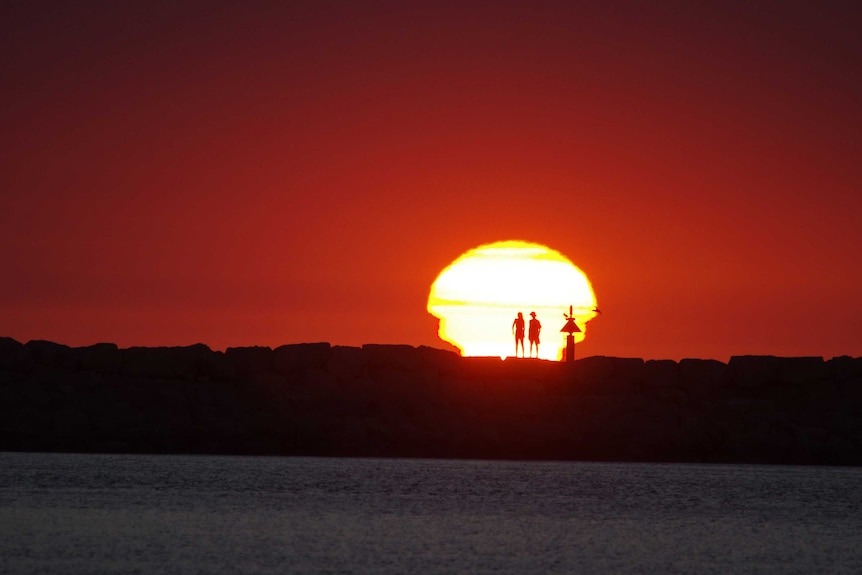 Two people silhouetted by a blazing yellow sun against a firey red backdrop.