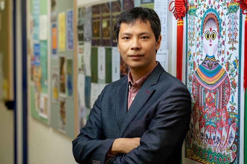 Zhouxiang Lu, with short black hair and wearing a suit jacket, stands against a wall with colourful print, and smiles slightly.
